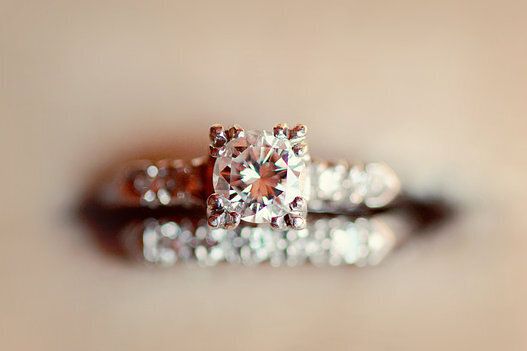 A Close-Up Shot Of The Engagement Ring Next To Blurred Wedding Ring