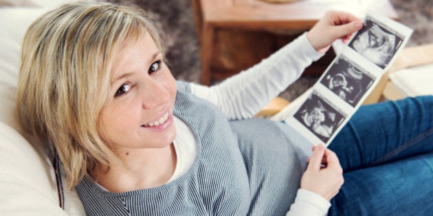 A pregnant woman sitting in a living room in a chair and holding an ultrasound image in the hands.