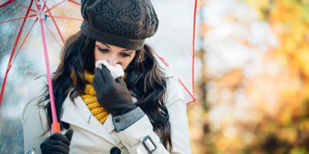 Sad woman with cold or flu blowing her nose with a tissue under autumn rain. Brunette female sneezing and wearing warm clothes against cold weather. Illness, depression and allergy concept.