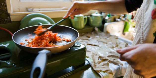Frying pan on old fashioned gas stove, with carrots and rice. Woman is stirring the ingredients. Wine leaves are drying on a dishtowel on the sink.