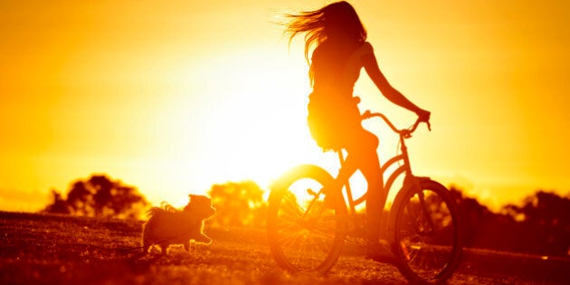 A silhouette of a young woman riding her bike with her dog running close behind.