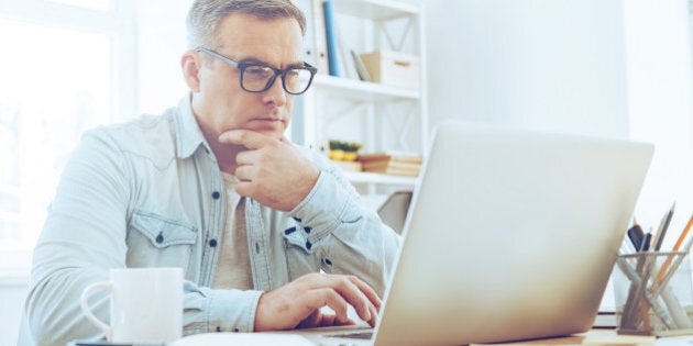 Thoughtful mature man looking at his laptop and keeping hand on chin while sitting at his working place