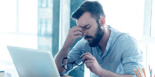 Frustrated young beard man massaging his nose and keeping eyes closed while sitting at his working place in office