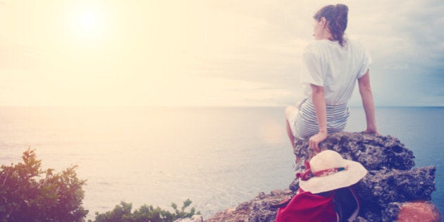 Young woman with hat and backpack sitting on a cliff and looking far away at the sea at sunset (intentional sun glare and vintage color)