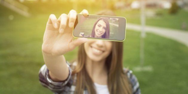 Beautiful young woman photographing herself with phone. Cute smiling young Caucasian teenage girl taking a selfie outdoors on sunny summer day.