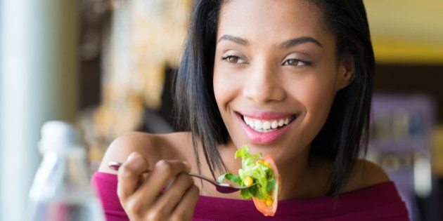 Closeup shot of young woman eating fresh salad at restaurant. Healthy african girl eating salad and looking away. Smiling young woman holding a forkful of salad. Health and diet concept. Woman ina a lunch break.