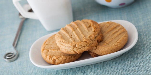 High Key image of 3 peanut butter cookies on a white oblong plate with a blue cloth along with a cup of tea and a polka dot teapot in the background.