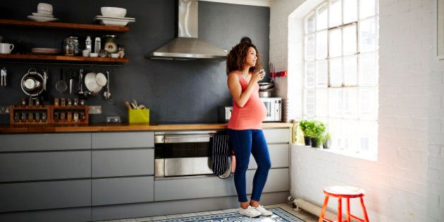 A pregnant woman drinks a cup of coffee whilst looking out of her modern kitchen on a sunny day