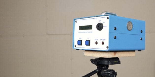 'A continuous radon monitor is mounted to a tripod in an unfinished house basement. A house is commonly tested for radon with a single use test kit. If initial readings are high or remediation work has begun a system like this would be used to determine a more accurate measurement over time. An added benefit of this unit is real time measurement. The display screen reads Avg. 3.5 pCi/l (picocuries per liter), the curie is a unit of radioactivity. The background is unfinished drywall with seams and screws.'