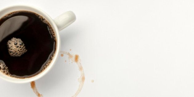 Overhead view of a white cup of freshly poured black coffee placed besides a ring of coffee that has stained the table top