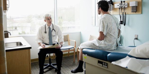 Male patient in gown sitting on exam table in discussion with doctor in exam room