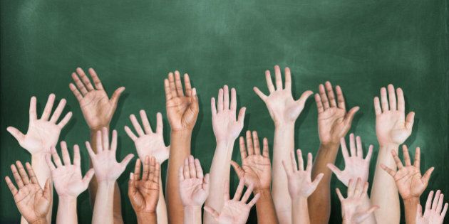 A multi-ethnic group of students' hands raised in front of a classroom blackboard. There are 18 hands raised, visible to the wrist or elbow, and seen from the side of the palm. The blackboard is dark green and has been freshly erased.