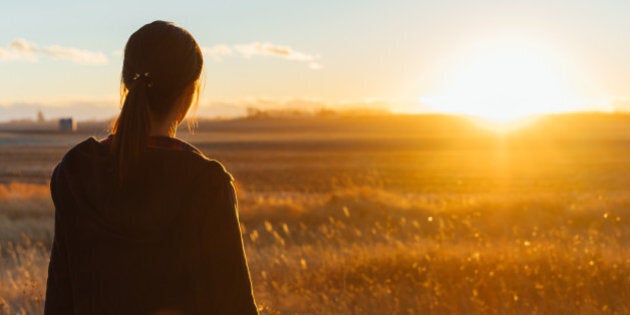 A Japanese woman is standing in a grassy field seeing the sunset in the distance.