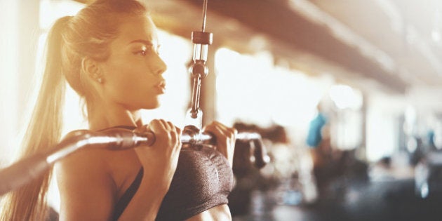 Closeup side view of an attractive brunette working out at a gym. She's using lat pull down machine with a reverse grip, the exercise that hits back muscles and arms. She's wearing tiny black sports top.Toned shot.