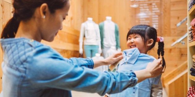 Pretty young mom fitting a shirt on the cheerful little girl at the kid's wear department in a fashion boutique