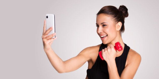Young mixed race woman making selfie while taking fitness exercises, standing on grey background