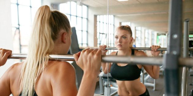 Muscular caucasian blond woman in a gym doing squats with a barbell on her shoulders while looking at mirror. Young woman working with weights at health club.