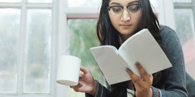 Indoor low angle image at day time in domestic room, near window of a beautiful, attractive Asian young woman reading a book while enjoying a mug of hot steaming coffee. One person, horizontal composition with selective focus and copy space.