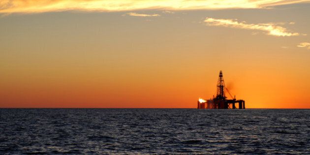 A wide angle view of an oil rig situated in the ocean exploring for oil and gas. The rig has a flame flaring on the side of the platform. The ocean is calm and the sun is about to set.