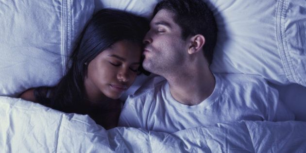 'Photo of an attractive young mixed-race couple lying on their bed, husband is kissing his wife goodnight on her forehead.'