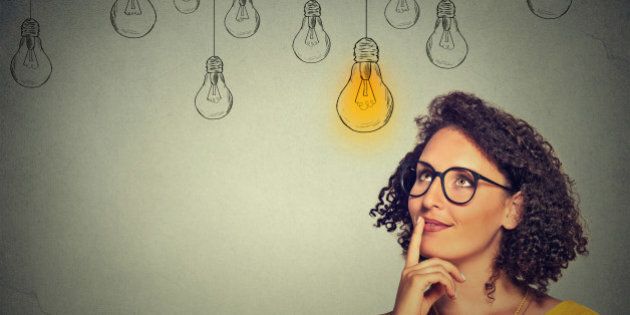 Portrait thinking woman in glasses looking up with light idea bulb above head isolated on gray wall background