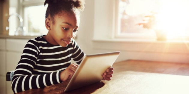 Young black girl with a fun afro hairstyle sitting at a table at home browsing the internet on a tablet computer with bright sun flare through the window alongside her