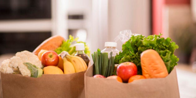 Bags full of groceries in paper sacks on a counter in a home kitchen. Food items include: apples, bread, lettuce, tangerines, bananas and a variety of other vegetables and food items. Domestic kitchen can be seen in background.