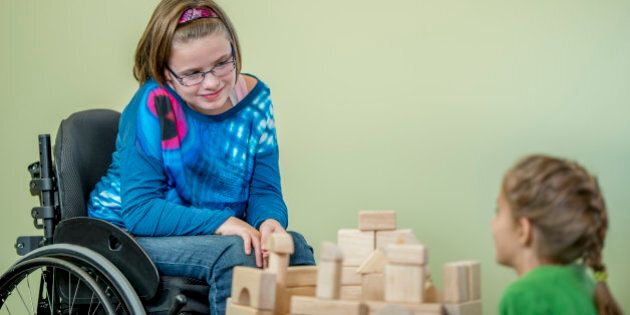 A young handicap girl is sitting in her wheelchair and is playing with other children - they are building towers using wooden blocks.