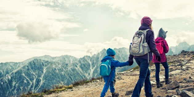 Mother and two kids hiking in mountains. The sky is cloudy. Everybody is wearing a backpacks and warm jackets.