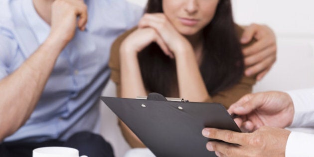 Close-up of thoughtful young couple sitting on the couch while man in formalwear pointing clipboard with document