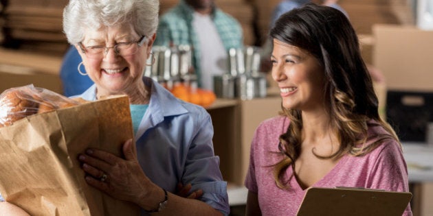 Cheerful Hispanic food bank manager helps a senior Caucasian woman. The older woman is carrying a bag full of groceries.