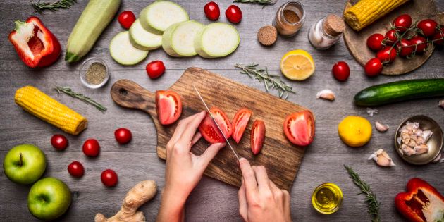 Female hand cut tomatoes on rustic kitchen table, around lie ingredients, vegetables, fruits, and spices, Healthy foods, cooking and vegetarian concept.