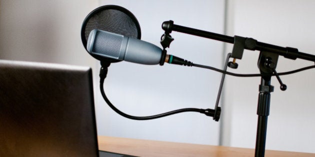 Horizontal image of audio equipment and a laptop sitting on a desk in a small sound room. The studio microphone is on a boom and has a pop filter in place. The laptop is open and ready to use.