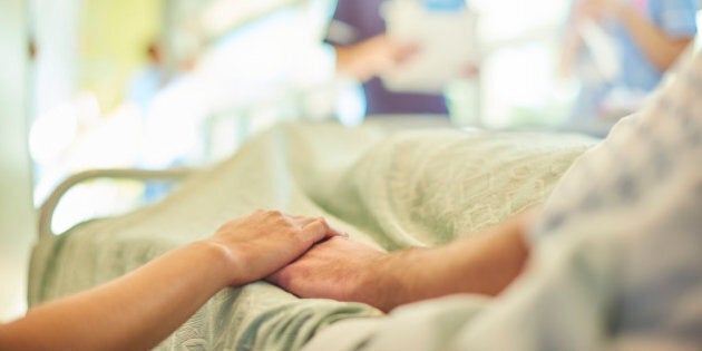 a hospital visitor's hand holds a patient's hand in bed of a hospital ward. In the blurred background a young nurse is chatting to the ward sister about the patient's care.