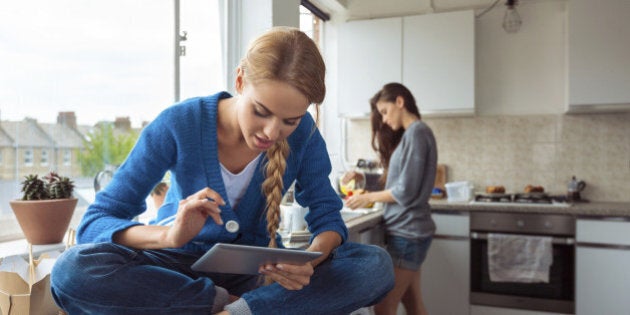 Roommates lifestyle. Young woman using a digital table while her friend washing dishes in the background.