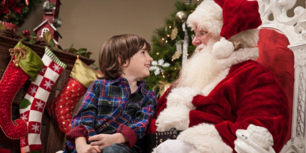 An excited young boy sits on Santa Claus' lap, looking up at him in awe, as they talk together.