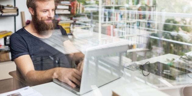 Young bearded man sitting on desk in his home office and working on laptop.