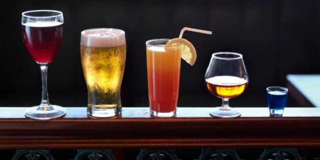 Standing on a banister, an assortment of various sizes and shapes of alcoholic drinks, ranging from shot, brandy, cocktail, beer and wine