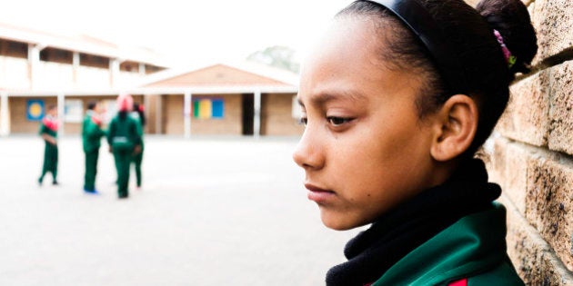 A sad school girl leans against the wall of a school courtyard. Other children play in the background.