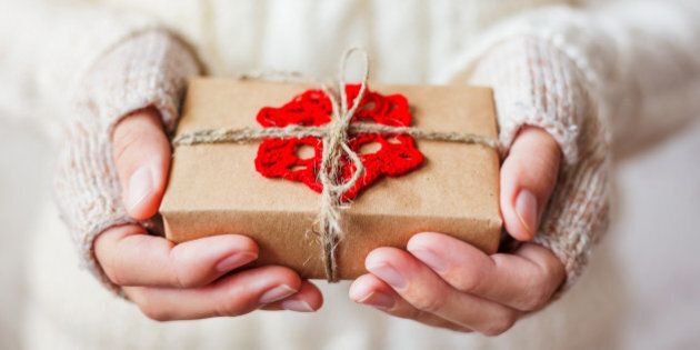 Woman in white knitted sweater and mitts holding a present. Gift is packed in craft paper with crocheted red snowflake. Example of DIY way to wrap a gift.