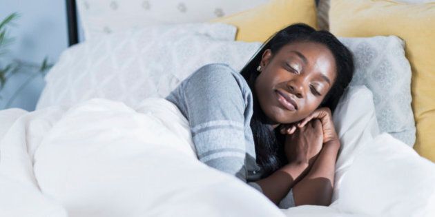 A young black woman lying in bed, on her side with her eyes closed, sleeping peacefully. It is morning or daytime, with sunlight on the white blankets. She is having a pleasant dream, with a smile on her face.