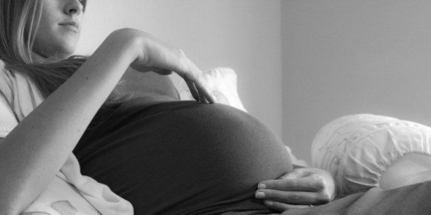 Pregnant woman in 3rd trimester reclines against pillows on a bed. Face partly visible as she gazes down with arms gently cradling her unborn baby.