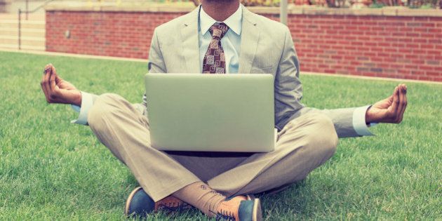 Young businessman with laptop meditating in lotus pose taking a deep breath outside corporate office. Business yoga and stress free environment. Peace of mind concept