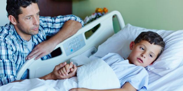 Tensed father looking at ill son lying in bed at hospital ward