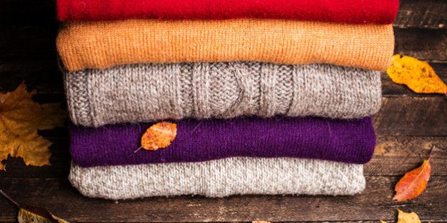 Pile of knitted winter clothes on wooden background covered with autumn leaves, knitwear, space for text. Stack of knitted sweaters and cardigans.