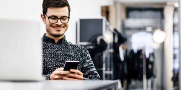 A young casual businessman wearing glasses is smiling at his smartphone in a bright modern office room. Clothes racks are seen in the background and a notebook in the front.