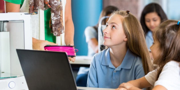 Elementary age Caucasian and Hispanic little girls are students in private school science and technology class. Students are using a laptop computer and learning 3D printing software. Girls are designing object to print with 3D printer. 3D printer is sitting on desk next to students. Diverse classmates are working on design projects in background.