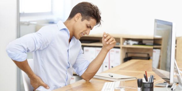 Shot of a businessman rubbing his aching back while sitting at his desk in front of his computer