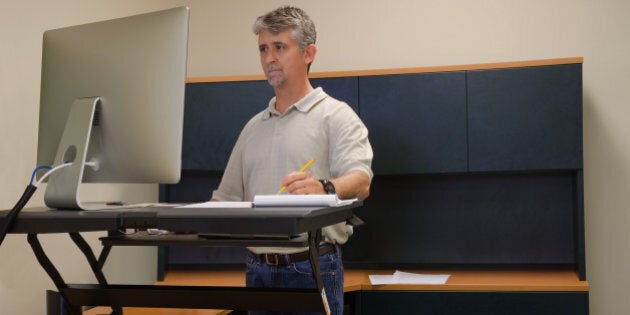 A man is working at a standup desk in an office where he works because standing is healthier than sitting all day. Live healthy, donât sit all day.