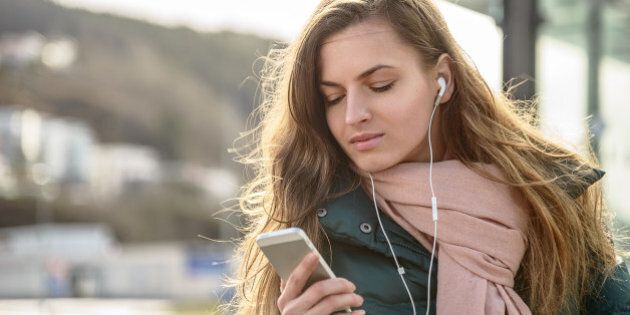 Young woman listening to music with headphones on her smart phone at a bus terminal/station during morning commute to work